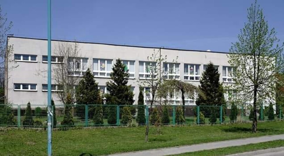 Jasny, dwupiętrowy budynek za zielonym ogrodzeniem i&nbsp;rzędem drzew iglastych.